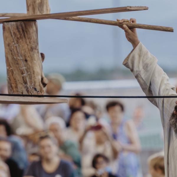 © Cia Nacho Flores L'artiste au premier plan tient dans une main une corde reliée à une structure en bois en équilibre, dont il tient une extrémité de l'autre main. Le public à l'arrière-plan est flou.