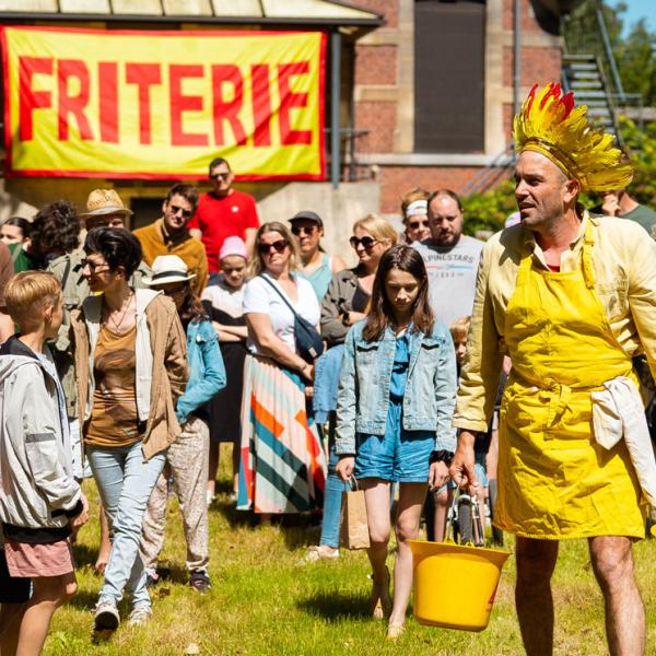 © Kalimba Un homme dans un jardin vêtu d'une couronne de plumes rouges et jaunes ainsi que d'une chemise et d'un tablier jaunes porte dans chaque main un seau jaune rempli de pommes de terres. Il est suivi par une trentaine de personnes de tout âges. Au fond, une grande bannière "FRITERIE" écrit rouge sur jaune est accrochée sur un bâtiment.