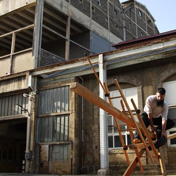 © Stéphane Rouaud L'artiste se tient en équilibre sur une construction en bois. En arrière-plan, un bâtiment industriel abandonné.