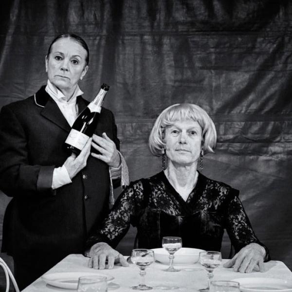 © Wilfried Thomas Photo en noir et blanc. Le comédien, habillé en bourgeoise, est assis à une table préparée pour un dîner. La comédienne est debout à gauche, habillée en majordome, et tient une bouteille de champagne.