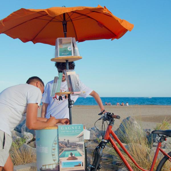 © Léa Lebrun Au bord de la plage, l'artiste se tient derrière son triporteur publicitaire. L'avant du vélo est constitué d'un parasol orange qui porte des flyers et d'un support en bois. Un homme est en train d'écrire, appuyé sur le plateau en bois.