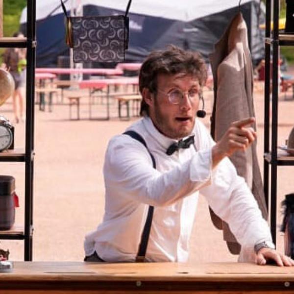 © Colas Lemaire Le comédien est debout derrière un haut bureau en bois, dehors. Habillé d'une chemise blanche, nœud papillon noir et bretelles, il porte un micro serre-tête et pointe le public du doigt. Derrière lui, des étagères sont remplies de bibelots, boîtes et objets de différentes tailles.