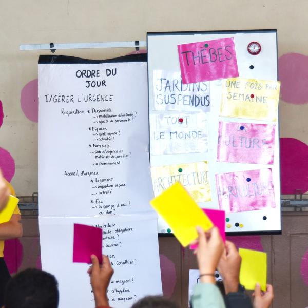 © Maxime Grimardias Au centre, deux panneaux blancs : l'un avec "L'ordre du jour", l'autre avec des pancartes colorées "Thèbes", "Jardins suspendus"... À gauche, la comédienne porte un t-shirt jaune et une pancarte jaune "Annulées". À droite, le comédien porte un t-shirt rose et une pancarte rose "Déplacées à l'extérieur". Le public brandit des cartons jaunes ou roses.