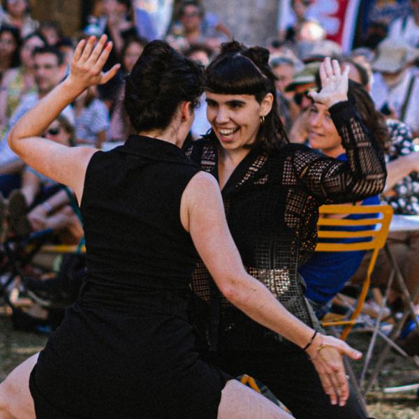 © Lucie Fournaise - Fournaise Factory Deux danseuses sont au milieu de personnes assises à des tables en extérieur. Elles se font face, accroupies et l'une sourit. Elles ont une main en l'air et l'autre en bas.