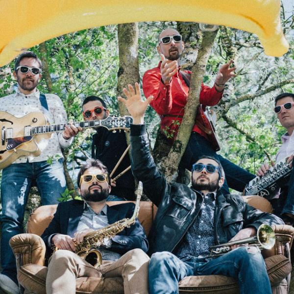 © Hélène Nugnes Dans un arbre, les six membres du groupes posent avec leurs instruments. Deux sont assis dans un canapé, les auyres sont debout ou posés sur des branches. Ils portent des lunettes de soleil et une banane gonflable flotte au-dessus du groupe.