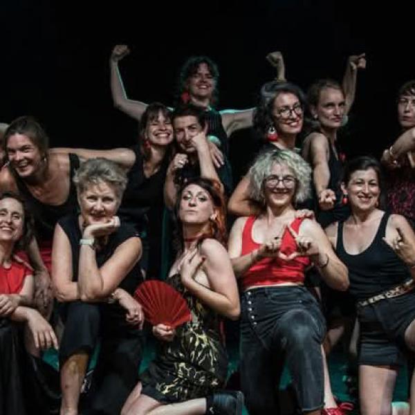 © Chorale de l'Ébranleuse Les membres de la chorale posent sur une scène en intérieur. La pièce est sombre. Elles portent du rouge et/ou du noir. Certaines font des signes avec les mains ou lèvent les bras.