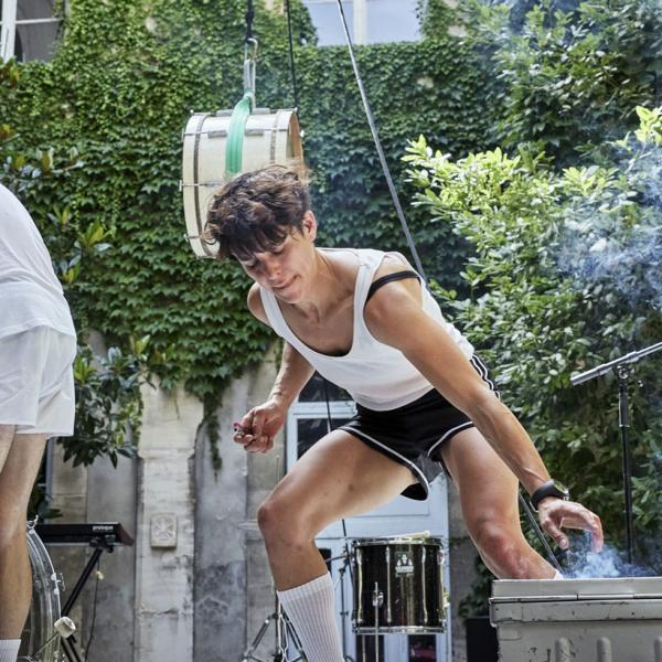 © Christophe Raynaud Delage Sur une scène dans une cour intérieure, une artiste allume un fumigène tandis qu'un musicien et une musicienne jouent de la batterie sur le côté. Un tom de batterie est accroché en l'air.