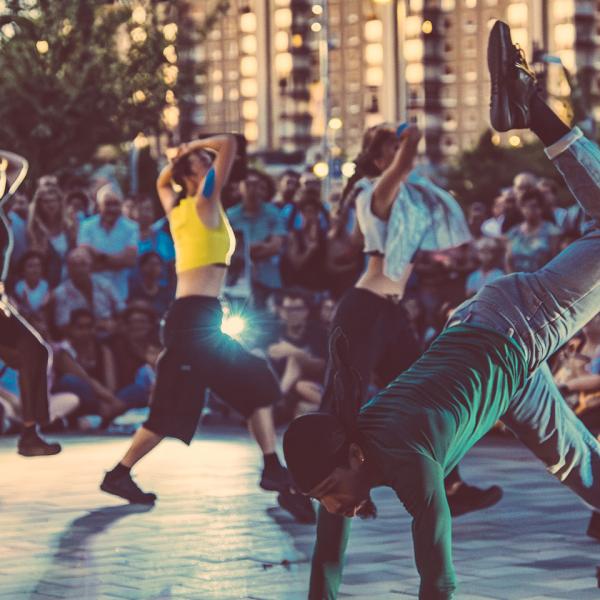 © Loïc Nys Une troupe de danseuses et danseurs en mouvement, encerclés par le public en extérieur. Un danseur à l'avant a deux mains et un pied à terre, et une jambe pliée en l'air. À l'arrière, cinq membres de la troupe avancent en diagonale en se couvrant la tête.