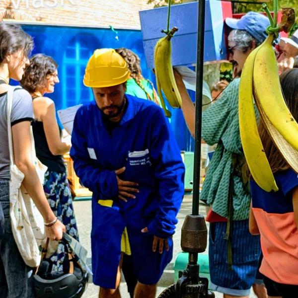 © Sandrine Velasco Des personnes dehors, un garçon tient une cannes à pêche avec des bananes au bout. Au milieu se tient un comédien avec une blouse bleue et un casque de chantier.