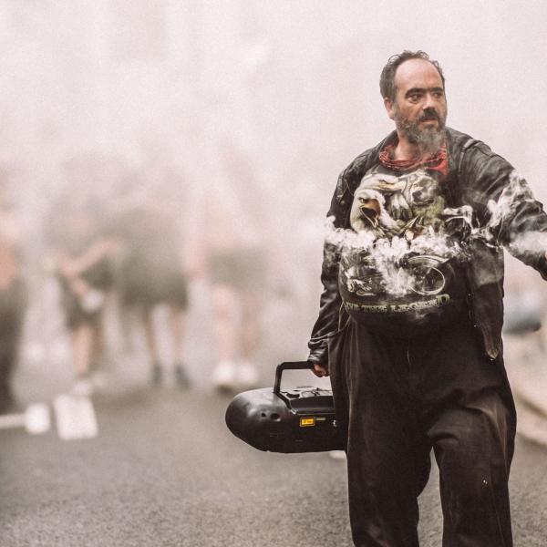 © Loïc Nys Un homme se tient au milieu de la rue. Il tient dans une main une grosse radio CD et dans l'autre un fumigène blanc. La rue est enfumée et un groupe de personne du public se tient à l'arrière, dans la fumée.