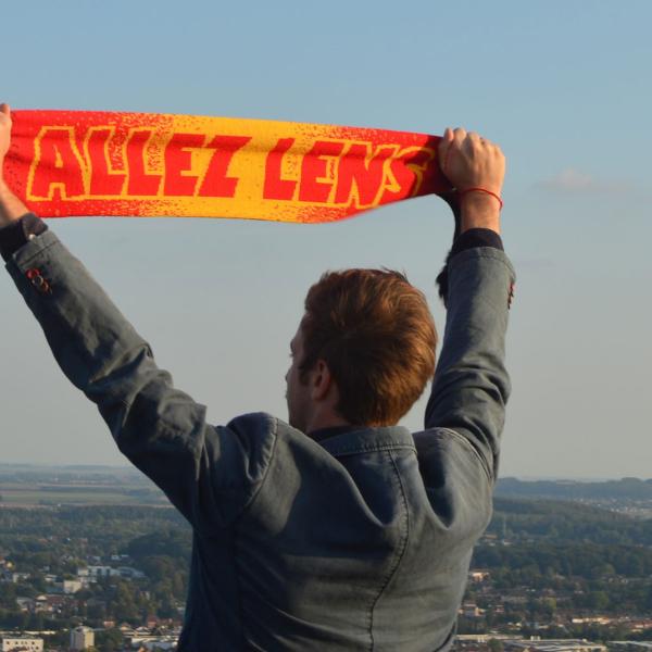 © Raphaëlle Viau Le comédien se tient de dos et brandit une écharpe de foot rouge et jaune "ALLEZ LENS". Il regarde l'horizon et les villes environnantes en contrebas.