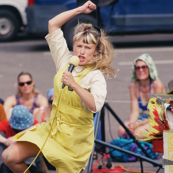 © Pierre Acobas La comédienne porte un tablier jaune et tient un micro dans sa main gauche. Elle semble en train de sauter, un bras en l'air et une jambe pliée. Derrière elle, un coupe-frites ambulant, décoré avec des motifs jaunes et l'inscription "Friterie". À l'arrière, le public est assis par terre, légèrement flouté.