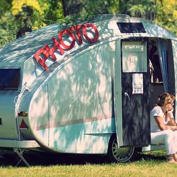© Clic-Omatic Caravane vintage dans une étendue d'herbe avec un lettrage "PHOTO" posé dessus. Une femme est assise sur le rebord de la porte.
