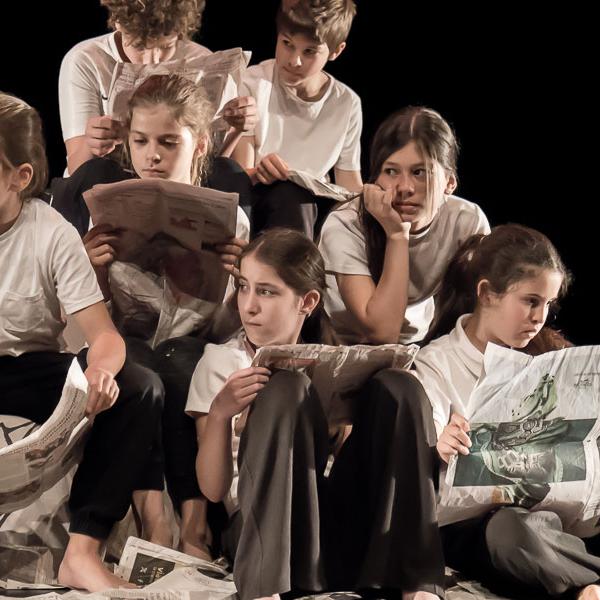 © XilloK Neufs enfants sont assis et assises sur scène et lisent des journaux. Iels portent un t-shirt blanc et un pantalon noir. Des journaux sont éparpillés sur le sol.