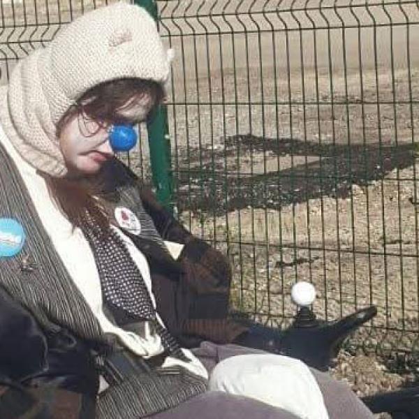 © Droits réservés Un clown en fauteuil roulant électrique porte une cagoule blanche avec des petites oreilles. Son visage est visible, peint en blanc et il porte un nez bleu en plastique. Au dos de son fauteuil sont accrochées des pancartes en carton.