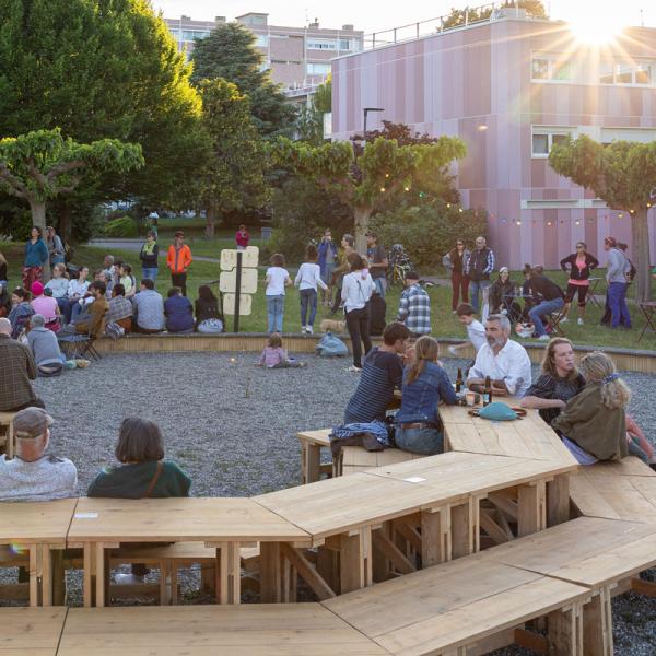© David Perpère Installation en bois de tables et bancs installés en octogone dans la Cité Rose/Résidence St. Agne de Ramonville. Des personnes sont assises et des enfants jouent autour pendant un concert.