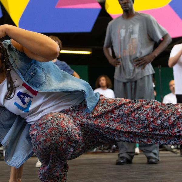 © Eduardo.B Une danseuse en plein mouvement de hip-hop. Elle a une main au sol, une jambe pliée et l'autre tendue vers le côté. Sa deuxième main couvre son front. Des personnes la soutiennent autour.