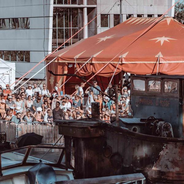 © Mathieu Lacout Photo d'une édition précédente du festival. Côté place du canal, les bateaux sont amarrés et le public se tient debout à côté d'un grand barnum rouge. L'entrée de l'espace est défini par une énorme arche en carton décorée de dessins avec écrit ARTO.